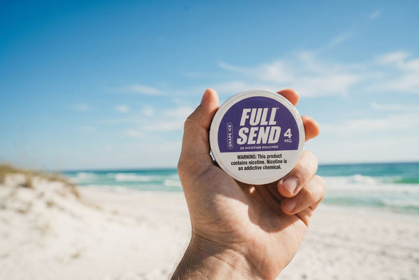Hand holding a 'Full Send 4MG' product container on a beach with blue sky and ocean.